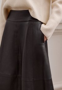 Schwarzer Lederrock mit hoher Taille, seitlichen Taschen und glatter Textur. Kombiniert mit einem hellbeigen, gerippten Pullover.