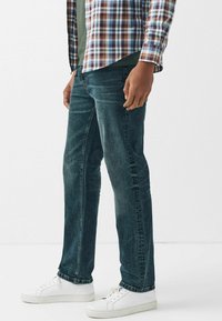 Tummansiniset farkut, joissa lievä haalistuma, suora lahjemalli ja minimaalinen ompelu, yhdistettynä valkoisiin tennareihin ja ruutupaitaan.