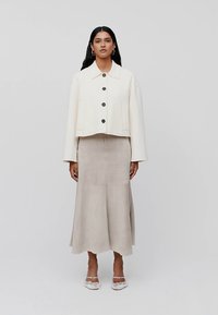 Krátká krémová bunda s velkými černými knoflíky a dvěma předními kapsami, kombinovaná s dlouhou světle šedou semišovou sukní. Čistý, minimalistický design.