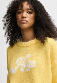 Maglione giallo lavorato a maglia con scollo rotondo e logo bianco. Tessuto testurizzato con una vestibilità ampia e polsini a coste.