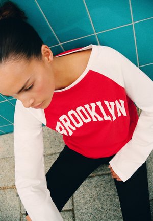 Jeune femme aux cheveux foncés attachés, portant un t-shirt rouge et blanc "Brooklyn", assise sur un sol carrelé gris devant un mur carrelé vert sarcelle.