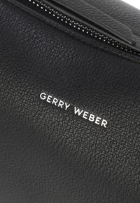 Sort tekstureret lædertaske med en toplynlås og "GERRY WEBER" logo i hvid, der understreger dens elegante design og holdbare materiale.