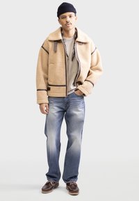 Braune Fleecejacke mit beigen Akzenten, Reißverschluss vorne und großem Kragen, kombiniert mit hellblauen Jeans und braunen Schuhen. Darunter ein schlichtes T-Shirt.