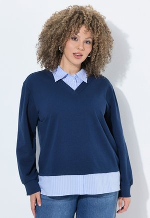 Kvinde med krøllet hår iført øreringe af ringe, lagdelt blå skjorte med marineblå sweater og blå jeans, stående mod en lys baggrund.