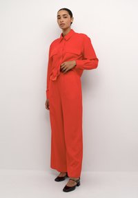 Rød bluse med sløjfedetaljer, parret med brede røde bukser. Model iført sorte hæle. Glat stof, minimalistisk design.