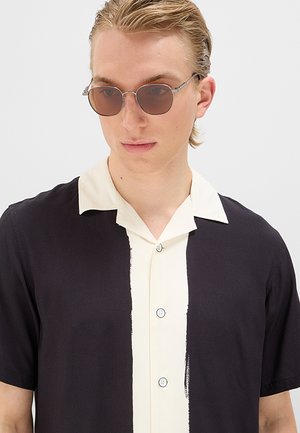 Giovane uomo che indossa occhiali da sole rotondi marroni e una camicia nera a maniche corte con una striscia verticale bianca e colletto.