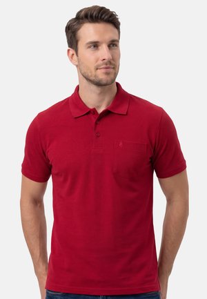 Mann trägt ein rotes Kurzarm-Poloshirt mit Kragen und Brusttasche, blickt leicht seitlich vor einem einfarbigen Hintergrund.