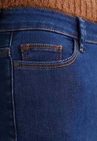 Dark blue denim jeans with a smooth texture, featuring a back pocket with contrasting orange stitching and silver hardware accents.