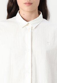 Camisa blanca de botones texturizada con cuello puntiagudo y un pequeño logotipo bordado en el pecho. Presenta una tela ligera y arrugada.
