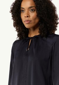 Granatowa bluzka z okrągłym dekoltem, z marszczeniami oraz zawiązywaniem. Materiał wydaje się gładki z lekko błyszczącym wykończeniem.
