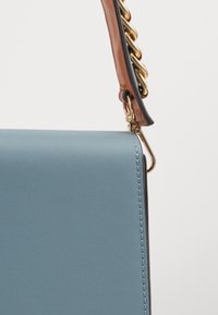 Blue leather handbag with a smooth texture, featuring a gold chain accent on a brown strap and visible stitching along the edges.