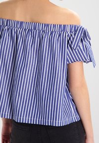 Bluzka na ramiączkach w niebiesko-białe pionowe paski. Wykonana z lekkiego materiału, z ozdobnymi węzłami na rękawach i luźnym krojem.