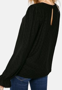 Blusa negra de manga larga con tela texturizada, hombros fruncidos y detalle de ojal en la espalda. Presenta puños con volantes y un corte holgado.