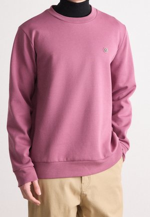 Personne portant un sweat-shirt mauve à manches longues sur un col roulé noir, associé à un pantalon beige clair, les mains partiellement visibles.