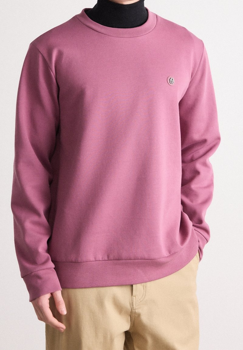 Personne portant un sweat-shirt mauve à manches longues sur un col roulé noir, associé à un pantalon beige clair, les mains partiellement visibles.