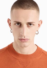 Nahaufnahme-Porträt einer Person mit kurzen Haaren, die einen gerippten orangefarbenen Pullover trägt. Sie hat silberne Creolenohrringe und eine neutrale Lippenfarbe.