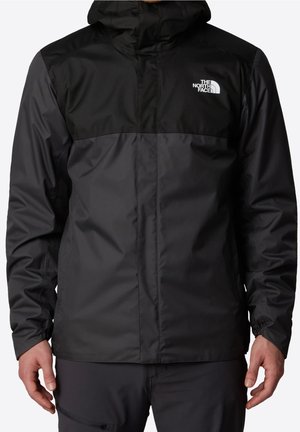 Mężczyzna ubrany w czarną i ciemnoszarą kurtkę z kapturem The North Face z przednim zamkiem, stoi z ramionami swobodnie opuszczonymi wzdłuż boków.