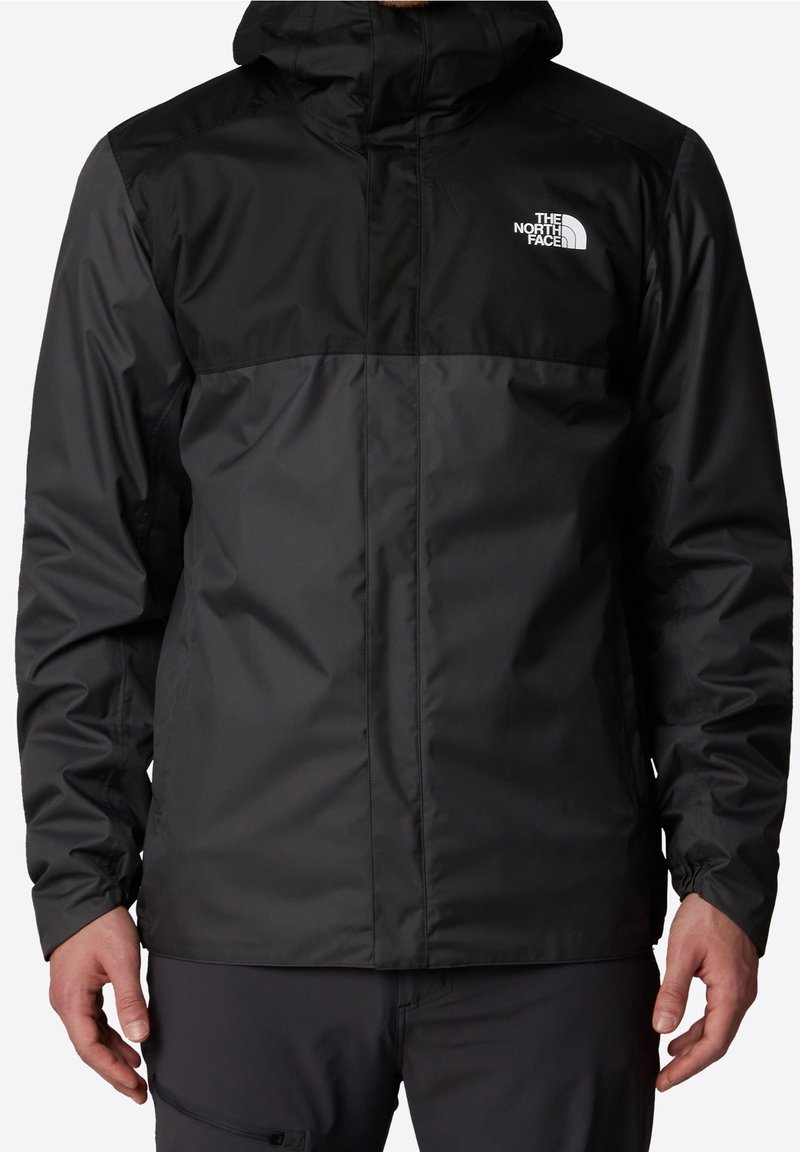 Homme portant une veste à capuche The North Face noire et gris foncé avec une fermeture éclair à l'avant, debout les bras détendus le long du corps.
