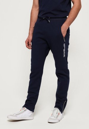 Mann trägt marineblaue Jogginghosen mit seitlichen Reißverschlusstaschen und weißen Turnschuhen, steht mit einer Hand in der Tasche.