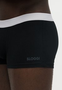 Primer plano de una persona que lleva bóxers negros con una cintura blanca y la palabra "SLOGGI" impresa cerca del muslo izquierdo.