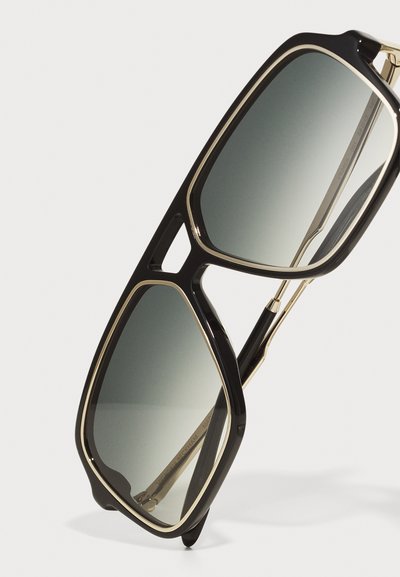 Svarta solglasögon med geometrisk design och guldaccenter. Gradientlinser som övergår från mörk till ljus. Tunna skalmar med detaljer.