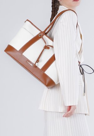 Mujer con traje blanco de rayas finas llevando un bolso tote blanco y marrón con correas de hombro sobre su hombro derecho.