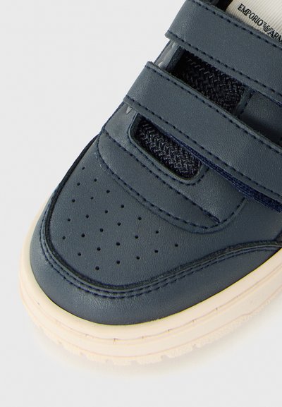 Mörkblå sneakers med perforerad tå, två kardborrband, svarta nätinslag och benvit texturerad sula på en vit bakgrund.