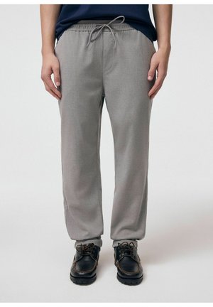 Osoba nosząca szare spodnie z sznurkiem i czarne sznurowane buty, stojąca na jednolitym jasnym tle.