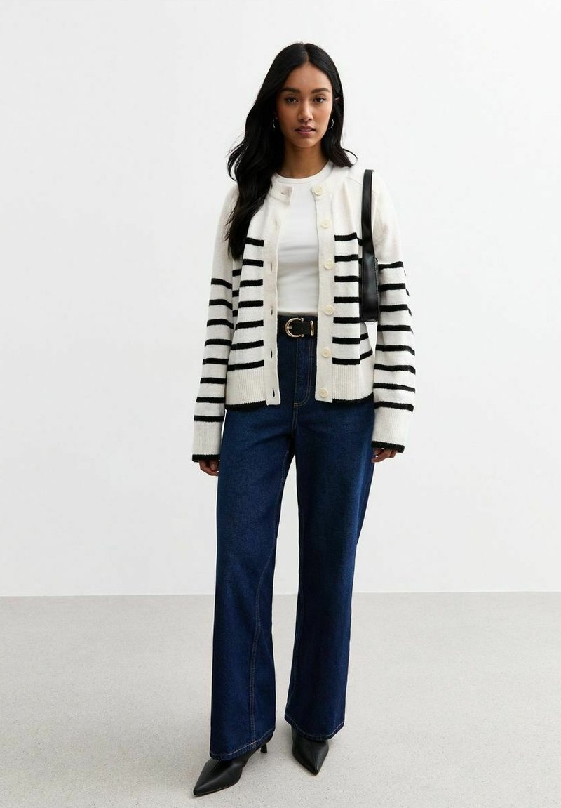 Gestreepte cardigan in wit en zwart, met knopen aan de voorkant, gedragen over een witte top, in combinatie met hoogzitende blauwe džins en zwarte puntige schoenen.