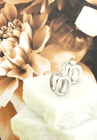 Silverfärgade metallörhängen med en slät, oval design, placerade på staplade, ljusfärgade stenblock mot en mjuk blommig bakgrund.