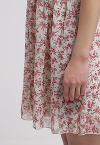 Květinové šaty z průsvitné látky, s bílým pozadím a malými růžovými růžemi a zelenými listy. Dlouhý, splývavý design s vrstvenou texturou.