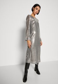 Srebrna sukienka z cekinami, z długimi rękawami, prostym krojem i okrągłym dekoltem. Połączona z czarnymi botkami do kolan. Minimalistyczny design i refleksyjna tekstura.