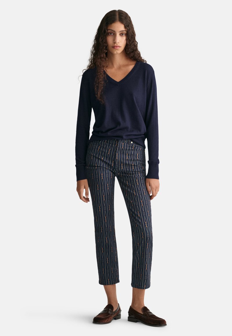 Marineblaue V-Ausschnitt-Strickjacke kombiniert mit gemusterten, knöchellangen Hosen mit vertikalen Streifen und geometrischen Akzenten. Dunkle Loafers runden das Outfit ab.