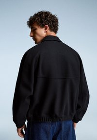 Veste en polaire noire avec col, coupe décontractée, poignets et ourlet élastiques. Présente une texture lisse et un détail sur le dos.