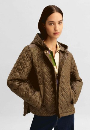 Giovane donna che indossa una giacca trapuntata marrone con cappuccio sopra una camicia a motivo, con le mani nelle tasche della giacca, su uno sfondo chiaro e semplice.