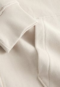 Gros plan sur un tissu de sweat-shirt beige montrant un poignet côtelé et une couture de poche avec des détails de couture visibles.