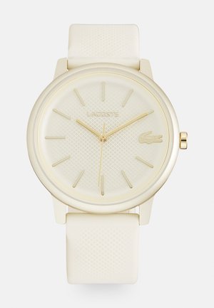 Světlý hodinky Lacoste s texturovaným ciferníkem, zlatými akcenty a silikonovým páskem. Kulatý design pouzdra v jemných bílých tónech.