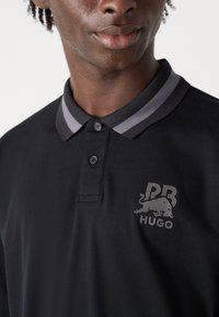 Zwarte polo met een grijsgestreepte kraag, een knoopsluiting met drie knopen en een logo met een gestileerde stier en initialen op de borst.