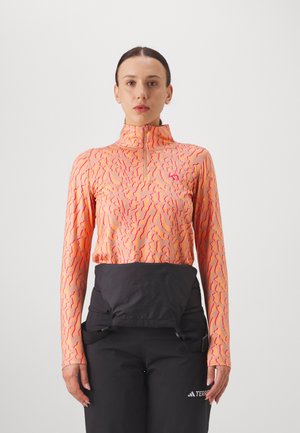 Sportieve lange mouw top in oranje met een roze golvend patroon, voorzien van een hoge kraag en een halve rits. Gekleed met zwarte high-waisted broeken.
