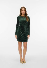 Ženska zeleno-večerja obleka iz žameta z dolgimi rokavi, prilagojena silhueta, nabran stranski detajl. Nošena z visokimi čeveljci z vzorcem, preprosta ogrlica.