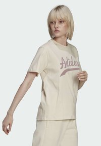 Bežinė medvilninė marškinėliai su ryškiu rožiniu "Adidas" logo priekyje, trumpomis rankovėmis ir laisvu siluetu, demonstruojami ant modelio.