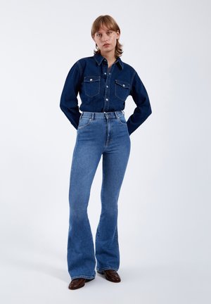 Młoda kobieta ubrana w ciemną dżinsową koszulę i wysokie, rozkloszowane niebieskie dżinsy, stoi z rękami za plecami na białym tle.