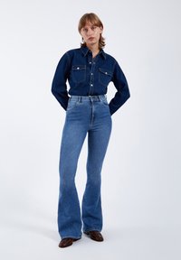 Camisa de mezclilla oscura con botones en la parte delantera y dos bolsillos en el pecho, combinada con jeans acampanados de cintura alta en azul claro y calzado marrón.