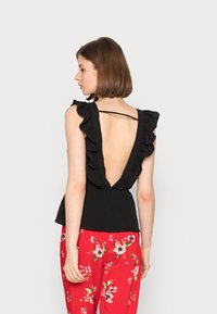 Top de alças preto com babados e design em V profundo nas costas, combinado com calças vermelhas florais. O tecido é suave e texturizado, realçando o estilo divertido.