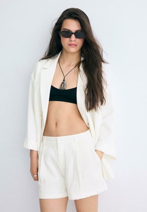 Žena s dlhými tmavými vlasmi, ktorá nosí čierne slnečné okuliare, čierny crop top, biely sako, biele krátke nohavice a náhrdelník s príveskom na jednoduchom pozadí.