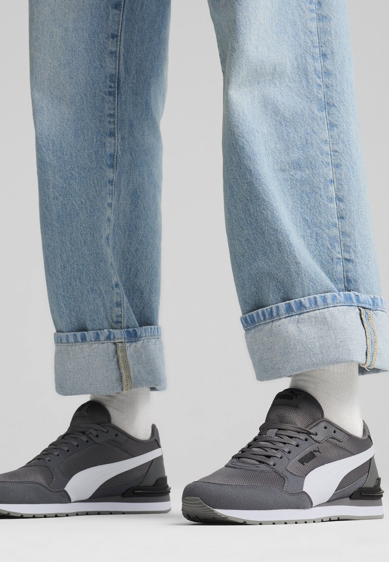 Personne portant un jean bleu clair retroussé et des baskets Puma grises avec des chaussettes blanches sur un fond uni.
