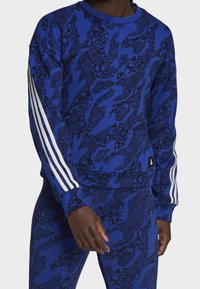 Sudadera azul con un patrón de camuflaje texturizado en azul oscuro, con tres rayas blancas en cada manga y un pequeño parche negro con el logo.