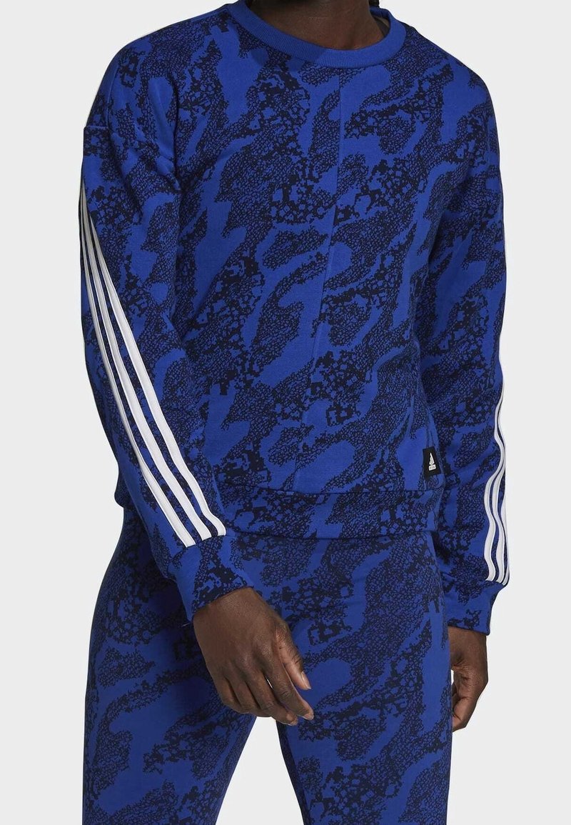 Sudadera azul con un patrón de camuflaje texturizado en azul oscuro, con tres rayas blancas en cada manga y un pequeño parche negro con el logo.