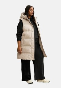 Chaleco de plumas color beige claro con capucha, con un diseño acolchado y cierres a presión. Lleva puesto sobre un conjunto negro con pantalones anchos y zapatillas.