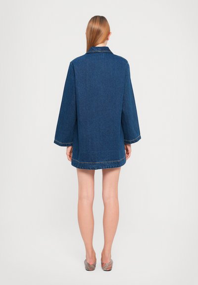 Dark blue denim jacket with a relaxed fit, featuring long sleeves, a collar, and hem stitching. Worn over a short garment, revealing legs.